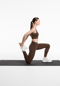 Brun atletisk crop top och leggings, modell som sträcker ut sig på en svart yogamatta, bärande vita sneakers, som visar upp det släta tyget och den figursydda designen.