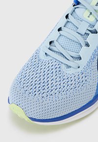 Zapatilla deportiva azul claro con parte superior de malla, patrón texturizado, cierre de cordones y acento en la suela verde. Punta redondeada y lengüeta acolchada.