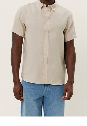 Hombre con camisa beige de manga corta abotonada y vaqueros de mezclilla azul claro, de pie frente a un fondo liso.