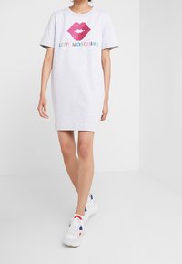 Kvinna klädd i en ljusgrå kortärmad klänning med en grafisk bild av rosa läppar och texten "LOVE MOSCHINO", tillsammans med vita sneakers med röda och blå detaljer.