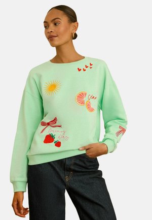 Donna con felpa verde menta decorata con cuori rossi, sole giallo, fette di agrumi rosa, fragole rosse e fiocchi rossi, abbinata a jeans scuri.