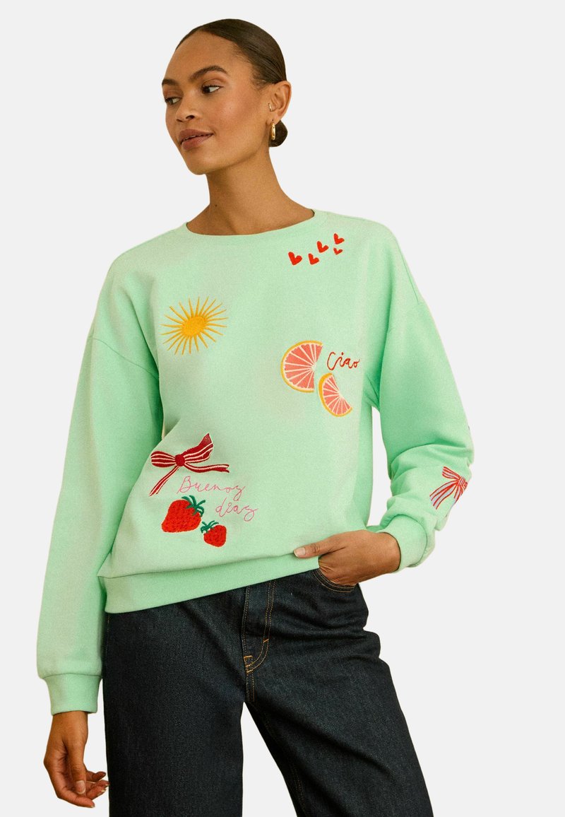 Kvinde iført mintgrøn sweatshirt med røde hjerter, gul sol, lyserøde citrus skiver, røde jordbær og røde sløjfer, kombineret med mørke jeans.