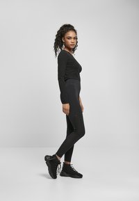 Haut noir à manches longues associé à des leggings noirs taille haute avec des panneaux latéraux texturés et des chaussures de sport noires, se tenant en profil.