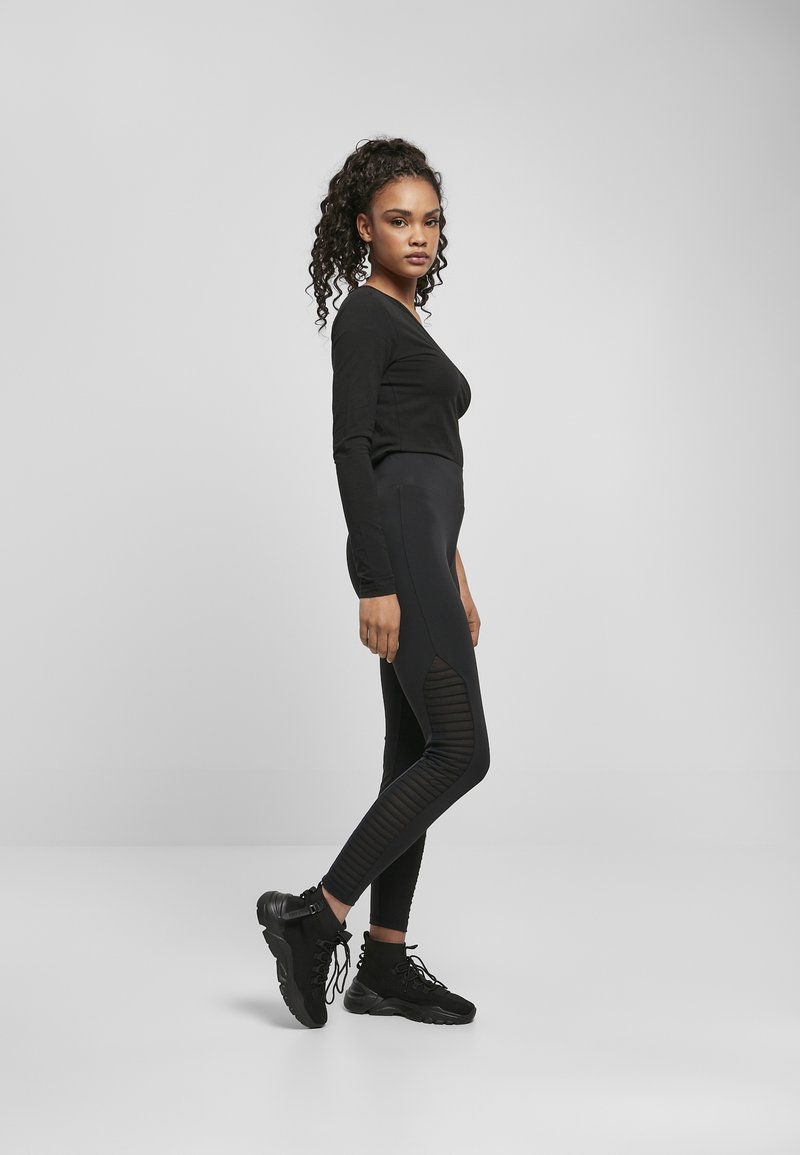 Haut noir à manches longues associé à des leggings noirs taille haute avec des panneaux latéraux texturés et des chaussures de sport noires, se tenant en profil.