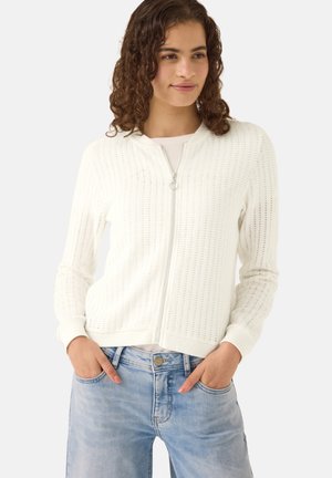 Frau mit lockigem Haar, die einen weißen gestrickten Reißverschluss-Pullover und hellblaue Jeans trägt, mit den Händen in den Taschen vor einem einfarbigen Hintergrund.