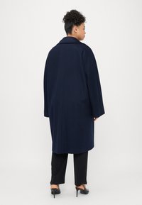 Cappotto blu in misto lana con colletto strutturato, spalle cadenti e vestibilità rilassata, abbinato a pantaloni neri e scarpe con tacco.