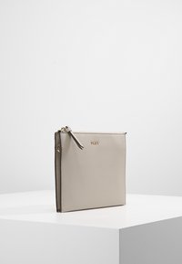 Pochette bracelet en cuir beige DKNY avec fermeture éclair et logo dorés, debout sur un socle blanc contre un fond uni.