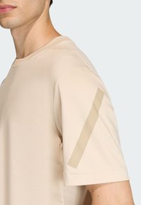 Camiseta de algodón beige con cuello redondo, que presenta un detalle brillante tipo pestaña en la manga izquierda. Textura suave y ajuste relajado.