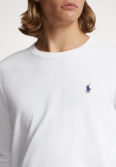 Balti ilgomis rankovėmis medvilniniai marškinėliai. Turi apvalų kaklą, aptemptą kirpimą ir mažą tamsiai mėlyną siuvinėtą logotipą ant krūtinės. Lygus paviršius.