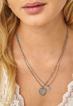 Close-up van de hals van een vrouw die twee zilveren hartvormige hangers draagt, waarvan één gegraveerd met "Amour", bovenop een wit kanten kledingstuk.