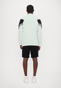 Camisola de fecho de correr verde menta com detalhes em preto e branco, corte solto e bainha canelada, combinada com calções pretos e sapatilhas brancas.