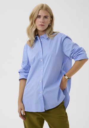 Žena s blond vlasmi oblečená v modrú pruhovanú košeľu na gombíky a olivovo zelené nohavice, s jednou rukou v vrecku a neutrálnym výrazom.