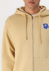 Lett beige zippered hettegenser med en myk tekstur. Har et blått blomstermotiv på venstre bryst og snorhette.