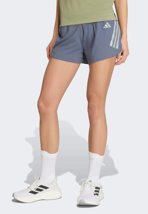 Nogi w niebieskich krótkich spodenkach Adidas z logo, białych skarpetkach Adidas i białych butach do biegania z czarnymi paskami, stojące na jasnym tle.