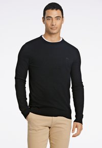 Maglione nero in maglia con maniche lunghe e polsini a coste, caratterizzato da una scollatura rotonda e un piccolo logo sul petto. Abbinato a pantaloni beige.