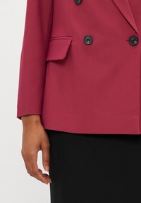 Blazer doppio petto bordeaux con bottoni neri, design strutturato, tasca a pattina, tessuto testurizzato e vestibilità leggermente allungata. Parte inferiore nera.