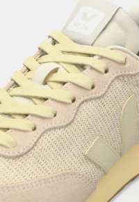 Μπεζ αθλητικό παπούτσι με υφασμάτινο ανώτερο μέρος με υφή και απαλές σουέντ λεπτομέρειες. Διαθέτει κίτρινα κορδόνια και ελαφριά λαστιχένια σόλα. Λογότυπο στη γλώσσα και στο πλάι.