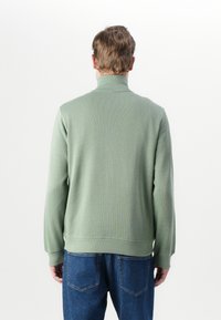 Groene geribbelde gebreide trui met een hoge kraag, voorzien van lange mouwen en een relaxte pasvorm, gecombineerd met blauwe spijkerbroek.