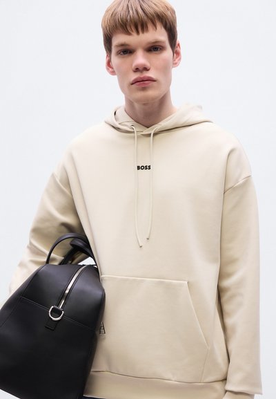 Sweat à capuche beige avec poche ventrale et cordon de serrage. Présente un logo noir "BOSS". Sac à dos noir avec fermeture éclair et ferrures argentées.