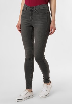 Person wearing fitted dark grey jeans and white sneakers standing on a plain light background.