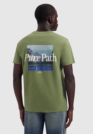 Man draagt een groen T-shirt met berg- en tennisbaanafbeelding en "Pure Path" tekst op de rug, gecombineerd met een blauwe spijkerbroek.