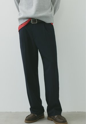 Persona che indossa pantaloni neri, maglione grigio, camicia rossa sotto e scarpe marroni, in piedi davanti a una parete chiara e semplice.