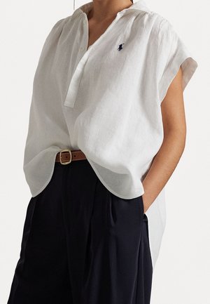 Personne portant une blouse blanche ample à manches courtes avec un petit logo marine, rentrée dans un pantalon noir taille haute avec une ceinture marron.
