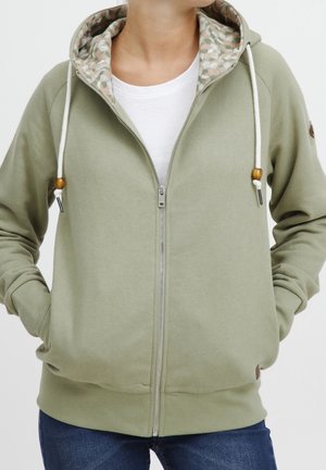 Personne portant un sweat à capuche zippé vert clair avec des cordons blancs et une doublure de capuche à motifs floraux, associé à un t-shirt blanc et un jean bleu.