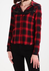 Chemise à manches longues à carreaux rouges et noirs, boutonnée, avec un col et un bord en dentelle noire le long de l'encolure. Texture de tissu lisse.