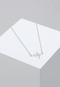 Collier en chaîne argentée avec pendentif en forme de battement de cœur présenté sur un piédestal géométrique blanc sur fond gris.