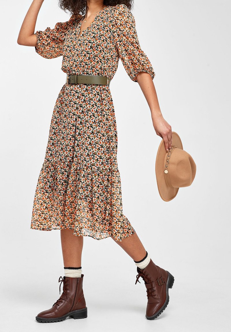Robe midi fleurie avec manches bouffantes, fleurs orange et blanches, ourlet à volants, associée à des bottines marron à lacets et un chapeau beige avec un détail chaîne.