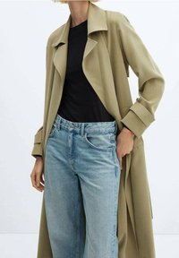 Cappotto trench beige con colletto ampio e maniche lunghe, indossato sopra una camicia nera e jeans azzurri chiari, caratterizzato da una vestibilità rilassata e design classico in denim.