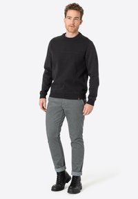 Zwarte sweatshirt met ronde hals en subtiele horizontale naaddetail, gecombineerd met grijze, opgerolde slim-fit broek en zwarte veterschoenen.