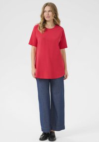 T-shirt in cotone rosso con maniche corte e orlo arrotondato, abbinata a pantaloni in denim scuro blu larghi e scarpe nere. Design semplice e vestibilità rilassata.