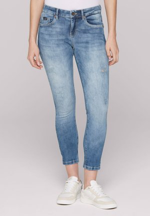 Woman wearing light blue skinny jeans and white sneakers, standing against a plain light gray background.