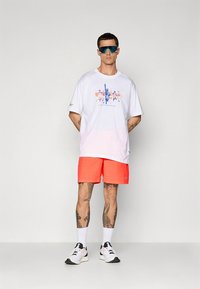 Balta T-krekls ar grafisku attēlu un gradienta efektu, kombinēts ar košām koraļļu krāsas šortām. Aksesuāri ietver atstarojošas saulesbrilles un sporta apavus.