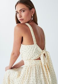 Žena s dugom kosom koja nosi kremastu haljinu bez rukava s crnim točkicama, s leđima vezanim velikom mašnom i zlatnim bisernim naušnicama.