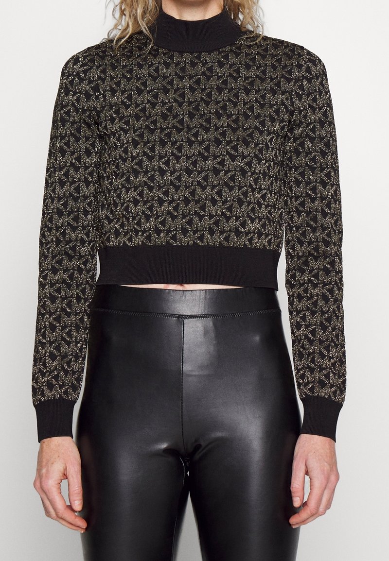 Maglione nero cropped con motivo geometrico dorato, collo alto a coste e polsini, abbinato a leggings neri in pelle a vita alta.