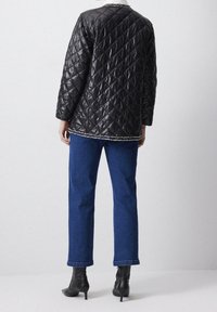 Person iført en sort quilted jakke med frynser, blå lige ben jeans og sorte hælede ankelstøvler, stående foran en ensfarvet baggrund.