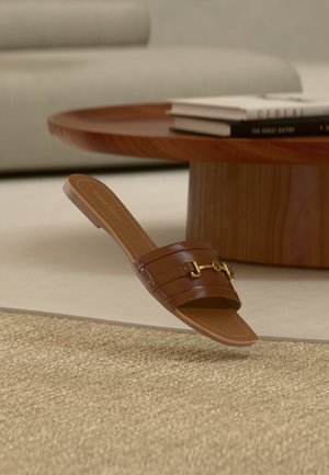 Sandale en cuir marron à enfiler avec détail boucle dorée, posée près d'une table basse ronde en bois et de livres empilés dans un salon minimaliste.