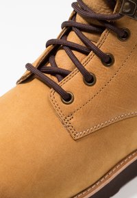 Bota de cuero marrón claro con cordones marrón oscuro pasados por ojales de latón y costuras detalladas en la parte superior y la suela.