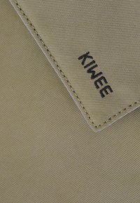 Tessuto verde oliva chiaro con una superficie testurizzata; presenta un logo nero "KIWEE" su un angolo ripiegato, bordato da una cucitura bianca.