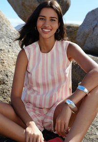 Young woman with dark hair wearing sleeveless pink and white striped top and shorts sitting on rocks outdoors, smiling at camera.