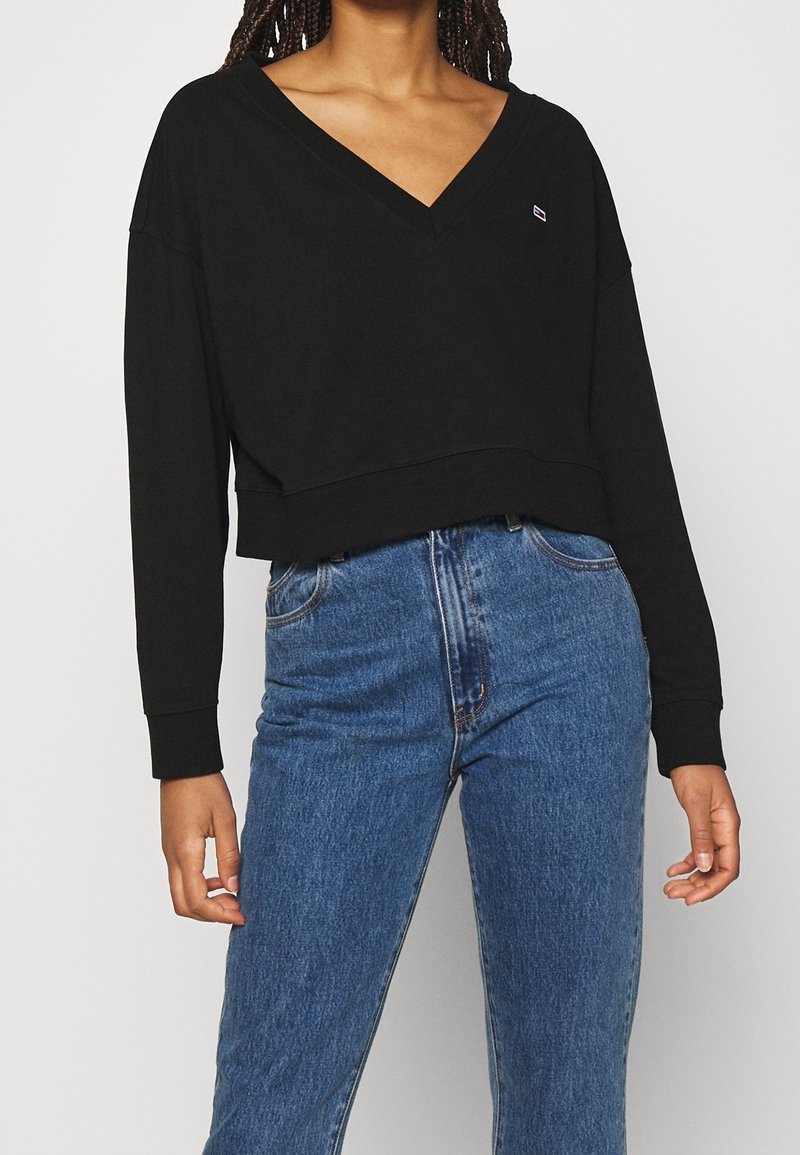 Femme portant un pull court noir à col en V et un jean bleu taille haute, debout devant un fond uni clair.
