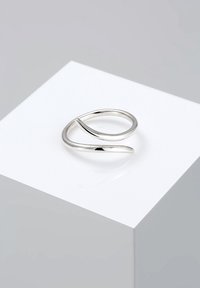 Bague ouverte minimaliste en argent avec un design lisse et courbé, présentée sur un cube blanc sur un fond gris.