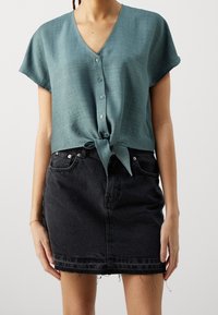 Frau, die eine türkisfarbene kurzärmelige, geknöpfte Bluse trägt, die an der Taille gebunden ist, und einen schwarzen Denim-Minirock mit ausgefranstem Saum.