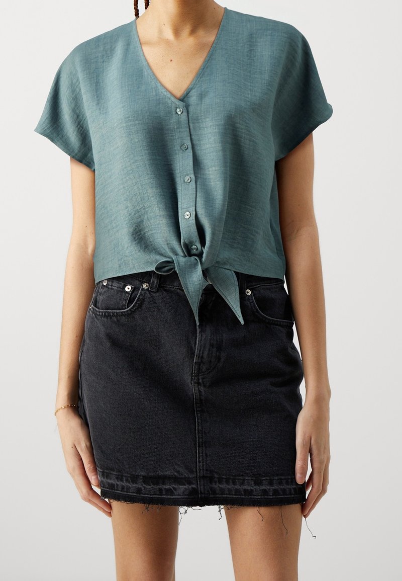 Frau, die eine türkisfarbene kurzärmelige, geknöpfte Bluse trägt, die an der Taille gebunden ist, und einen schwarzen Denim-Minirock mit ausgefranstem Saum.