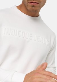 Hvid sweatshirt med forhøjet "INDICODE JEANS" tekst, rund halsudskæring og ribbede manchetter. Blødt stof med en glat tekstur.