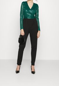 Top en sequins teal avec un décolleté en V et des manches longues, associé à un pantalon noir fuselé et un petit sac à main noir en relief crocodile. Escarpins noirs.