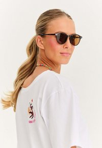 Žena s blond ohonem, která má na sobě tmavé sluneční brýle, bílé tričko s barevným grafickým potiskem na zádech a korálkový náhrdelník.